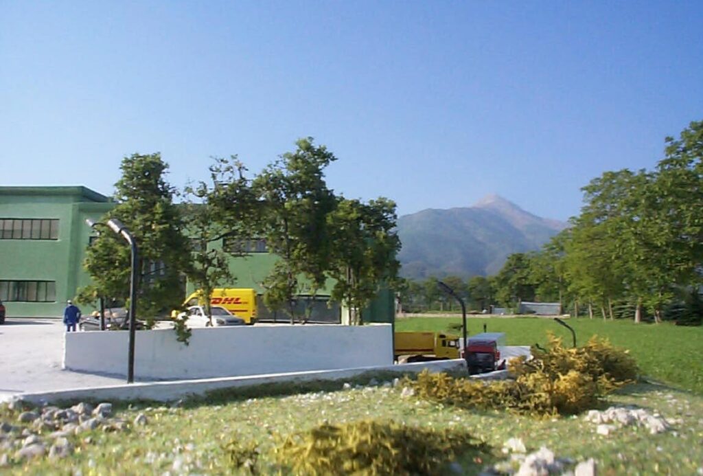 Edificio verde con furgone giallo in un paesaggio rurale, alberi e montagne sullo sfondo.