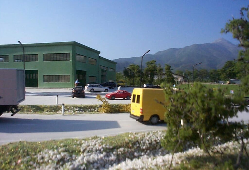 Edificio industriale verde con veicoli parcheggiati, sfondo di montagna e cielo blu.