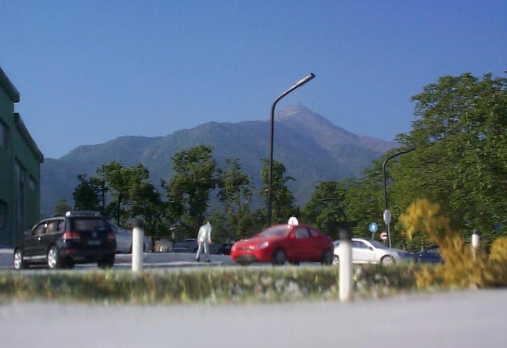 Auto in sosta su strada alberata con montagna sullo sfondo, cielo sereno e lampioni.