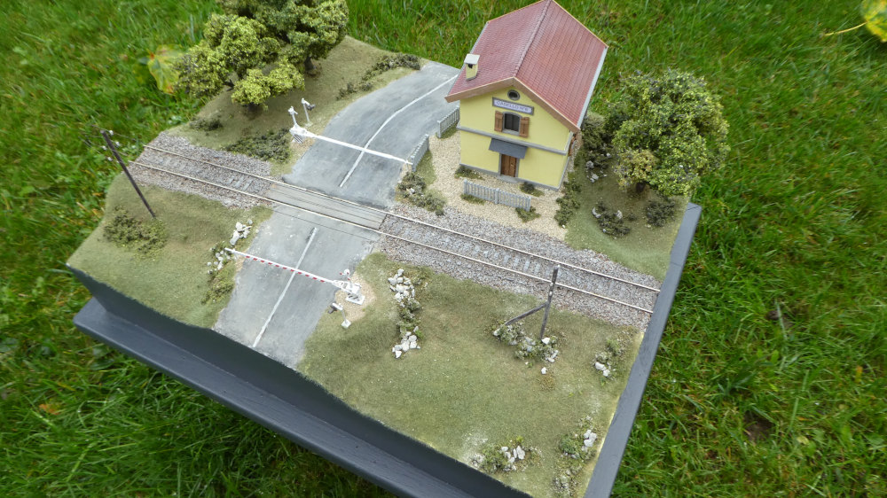 Modello in scala di un passaggio a livello ferroviario con casa, alberi e strada su prato verde.