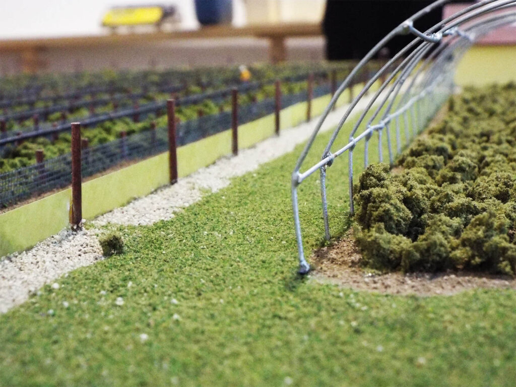 Modellino di una serra con struttura metallica e vegetazione verde, su un terreno erboso con ghiaia.