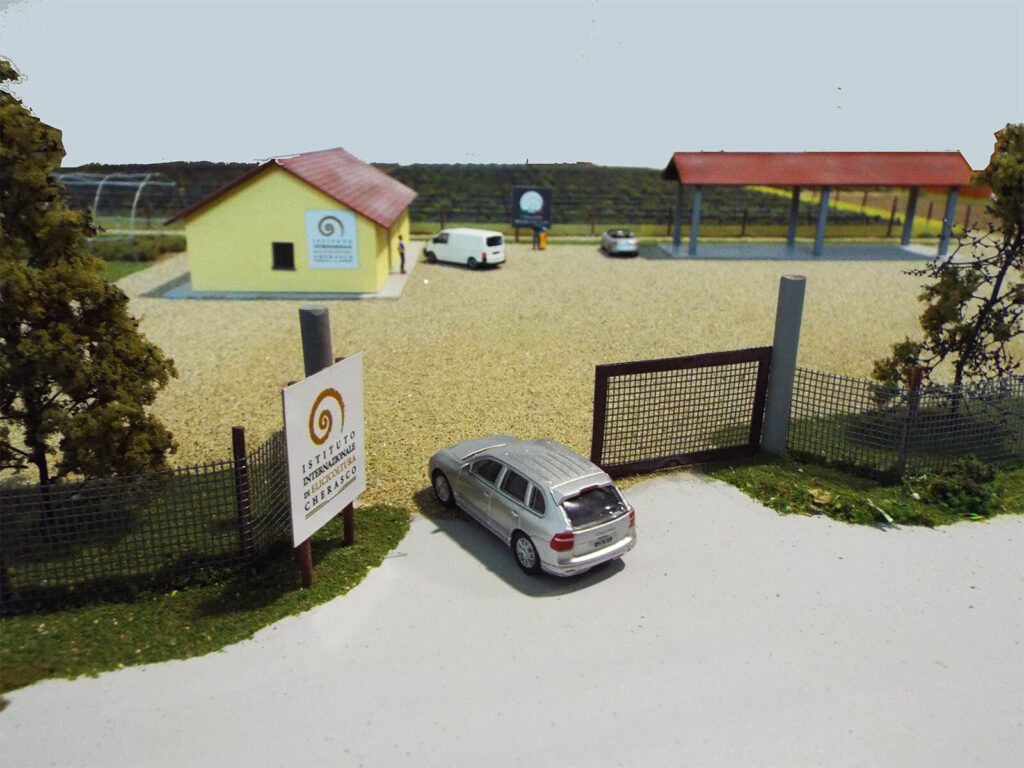 Modellino di un edificio giallo con tetto rosso, parcheggio e cartello. Auto argento visibile davanti al cancello.